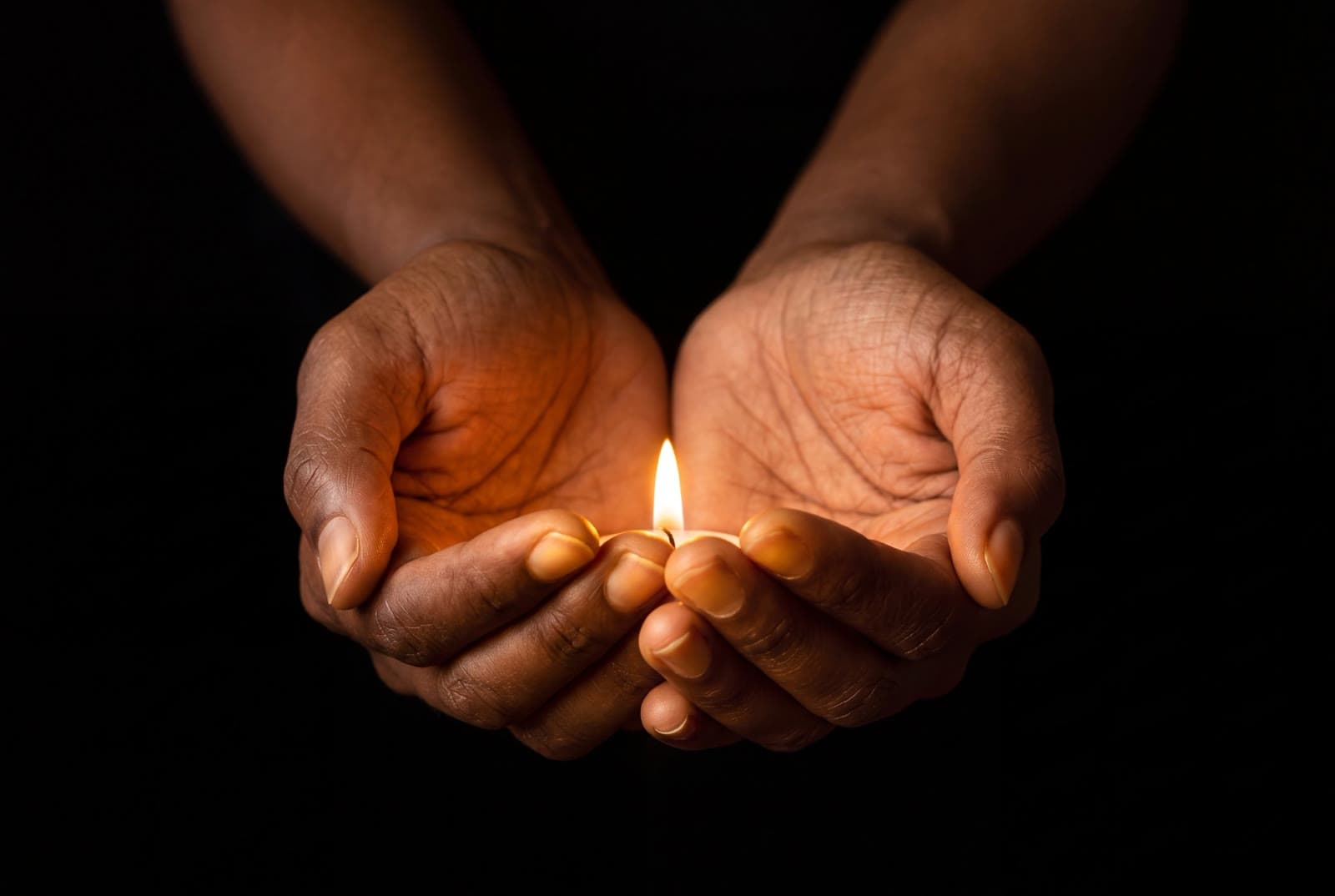 Two deep brown hands cupping a single controlled flame against a black background